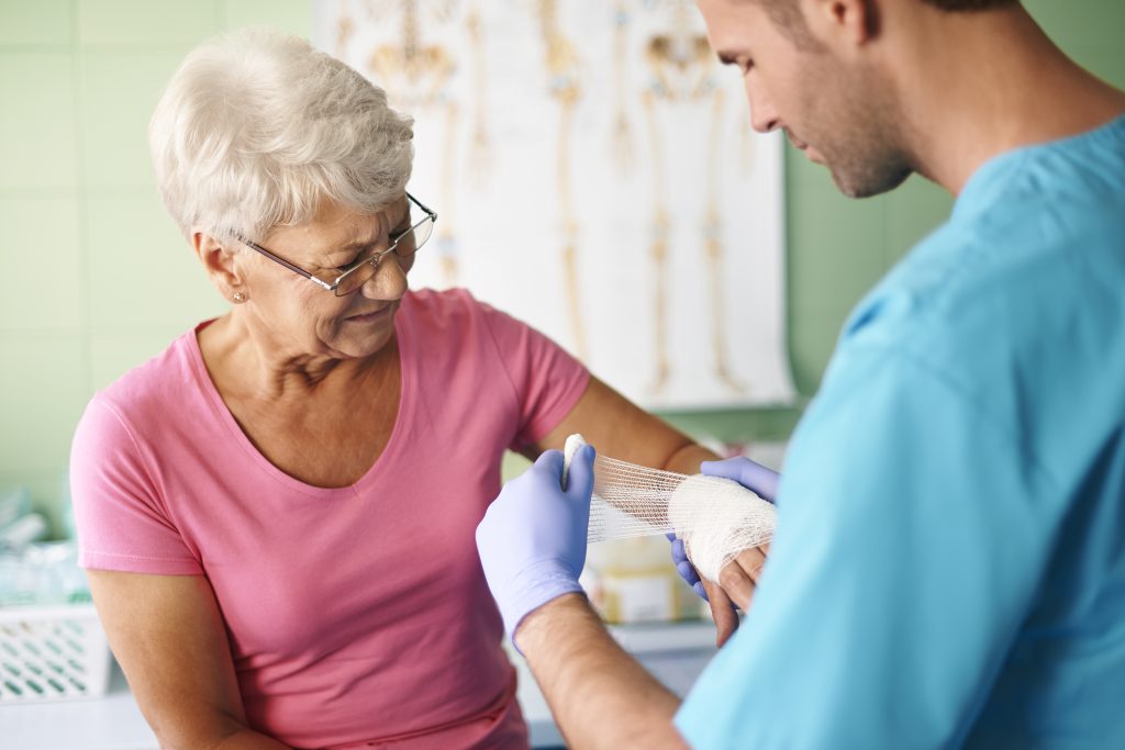 An elderly English woman with short hair inside her home is receiving medical help and bandage treatment on her left hand from a male professional caregiver as part of geriatric care at home as immediate help.