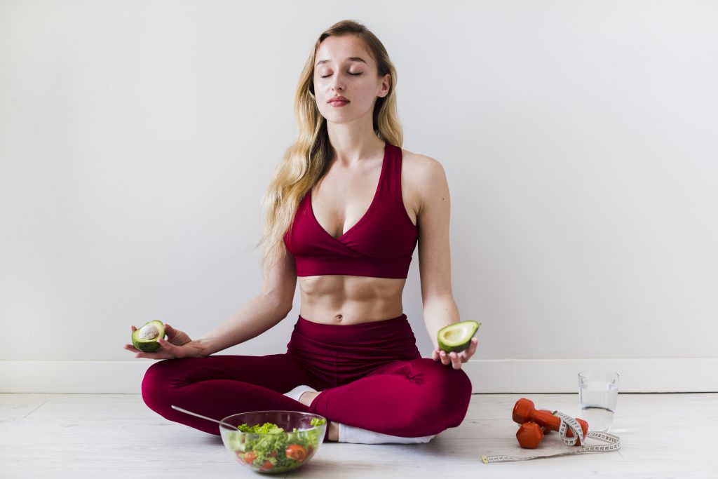 lifestyle changes-a young women mediatating in her yoga pants keeping her eyes closed while holding healthy fruits in her hands showcasing healthy lifestyle actions for young female.