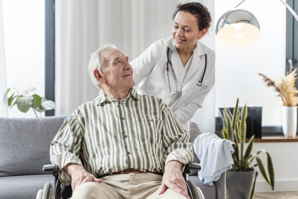 A white elite man cheerfully sitting on his wheel chair inside his living room and receiving professional healthcare from a smiling compassionate doctor as part of home health care services. Professional nursing care at home in Dubai